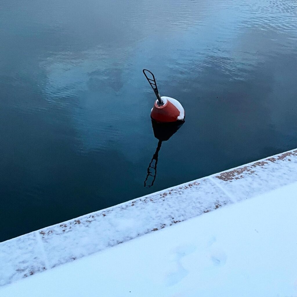 Red buoy partly covered in snow, floating near a wintery shore.
